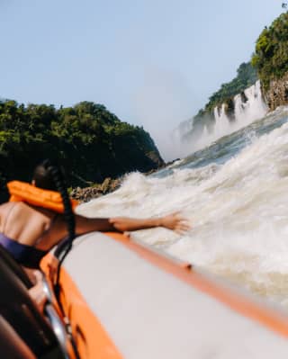 A woman in a lifejacket reaches from a Macuco Safari boat to feel the river spray, seen from behind with the falls up ahead.