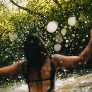 A spray of droplets adds a magical touch to an image of a woman playing in a river with verdant banks, seen from behind.