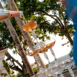 Angled image of a three-tier pyramid of coupe glasses, with pink champagne pouring into the top one, at an al fresco event.
