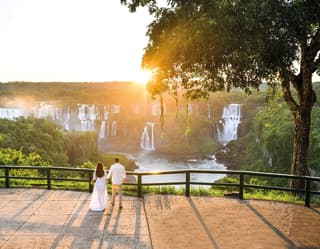 A man in white shirt and woman in long white dress hold hands gazing at the cascades of Iguassu Falls