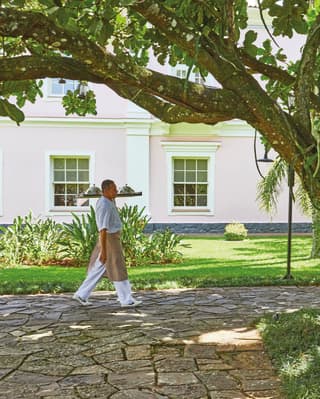 A male staff member in whites and a beige apron carries a tray with cloche-covered plates through the fern-filled gardens.