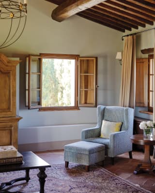 A pale blue wing backed chair sits near the open window of a traditional Tuscan sitting room with terracotta floor