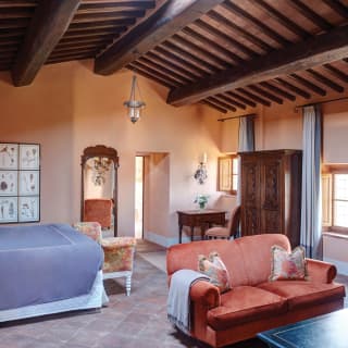 A vintage room with exposed beams and terracotta floor features a sienna sofa with blanket, drapes and bedlinen in blue-grey