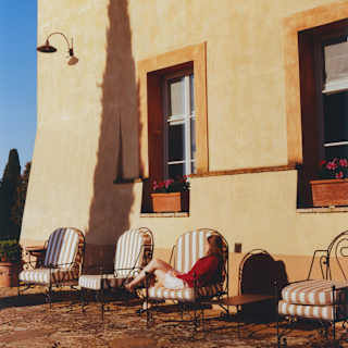 A woman relaxes in one of four patio chairs by a sunny exterior wall brushed by the tall shadow of a cypress tree off camera.