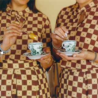 Two women in identical brown and white-checked dresses stir coffee in delicate cups, side by side, seen close-up.