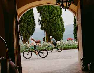Tilted view of a man and woman as they cycle past an arched doorway with a backdrop of cypress trees, seen from the interior.