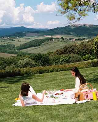 Two women relax on a white picnic blanket laid out on a gentle grass slope with a hamper of food and rolling hill views.