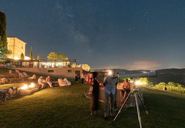 Some guests sit on the amphitheatre steps with lamps, while others stand by a telescope below a night sky sprayed with stars.