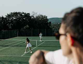 A couple play tennis on a grass court watched by a man in shades in soft-focus foreground, resting an elbow on a wire fence.