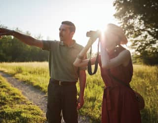 A young man dressed in gamekeeper green points into the distance. Beside him a woman guides her binoculars to his line
