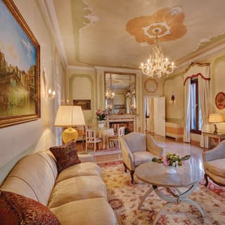 A palatial lounge is filled with ivory silk sofas, cream and red floral rugs and an Old Master painting of the Rialto Bridge