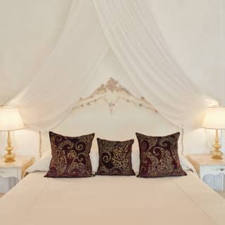 Gold and burgundy cushions top a king bed. Hand painted flowers on a Rococo headboard peek out from a voile bed crown