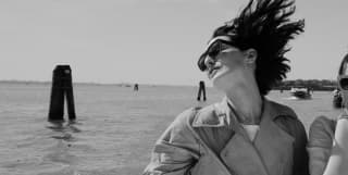 A chic young woman in a classic trench coat and sunglasses allows the wind to catch her hair aboard a boat on the lagoon