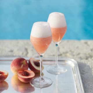 Two classic Bellini cocktails stand on a silver tray with freshly cut white peaches. Behind, the pool is a cool azure