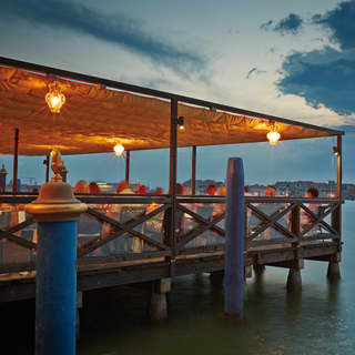 Diners sit under a canvas awning, lit by warm yellow glass globes at dusk. Below their wooden terrace, canal waters swirl