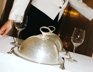 A waiter pushes a table for two, with wineglasses, cutlery and plates covered with silver cloches, in a room service detail.