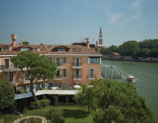 Pines cast pools of shade across green lawns. The private launch approaches the hotel dock. Pink walls stand majestic