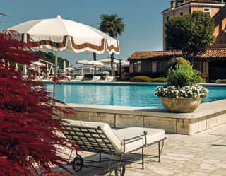 A cloudless sky mirrors the blue pool. In the foreground a red fern. Behind the pool, majestic pinks walls and terracotta roofs