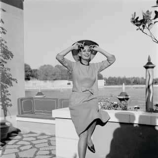 A black and white image of Italian actress, Sophia Loren, smiling as she sits on a low wall in grounds of the Hotel Cipriani