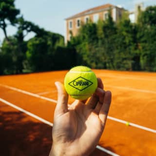 A man's hand holds a Cipriani-branded yellow tennis ball, with the clay-surfaced court and hotel in the background.