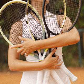 A woman, seen from the neck down, uses both arms to hold three tennis racquets flat against her chest, seen on a clay court.
