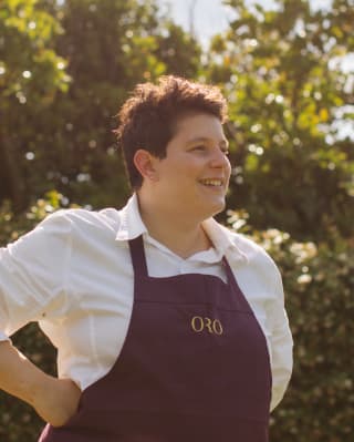 Oro Head Chef Vania Ghedini, with short dark hair, wearing a branded apron, smiles in a portrait taken in the hotel gardens.