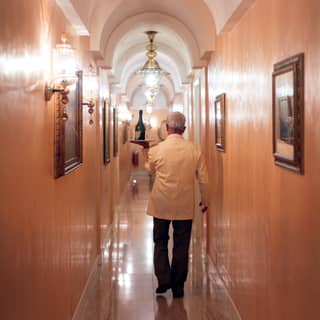 A waiter carries aloft a silver tray of wine and glasses for room service, through a vaulted corridor lit by Murano glass lights