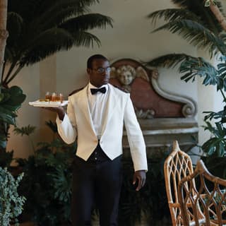 Bar waiter dressed in white tux and bow-tie carrying a tray of two whisky glasses