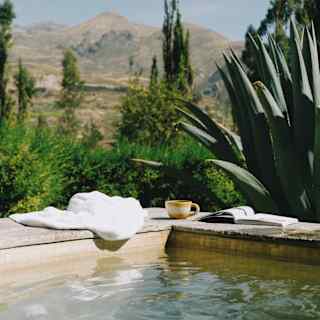 A white towel, mug and book rest on the corner edges of a pool in an eye-level view from the water, capturing hills beyond.