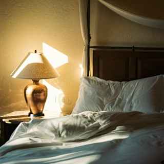 Sunlight dances on a wall behind a bed in a view of the bedside with a table and lamp, and white rumpled bedding.