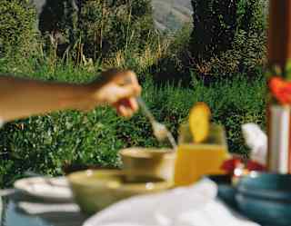In soft-focus foreground, a woman stirs her coffee at an outdoor breakfast table, with a backdrop of garden trees and hills.