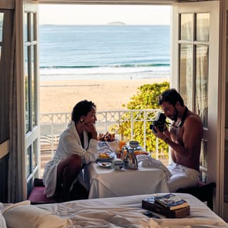 A woman sitting at the breakfast table facing her partner while he takes photographs of her with a vintage camera