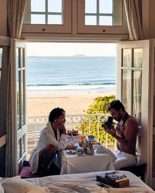 A woman sitting at the breakfast table facing her partner while he takes photographs of her with a vintage camera