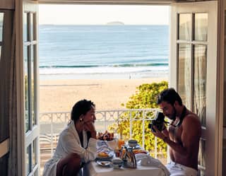 A woman sitting at the breakfast table facing her partner while he takes photographs of her with a vintage camera