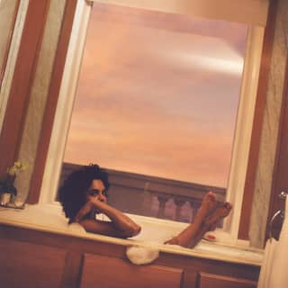 A woman with dark curly hair soaks in a foam bath with her feet up on the sill of the bathroom window, framing the sunset.