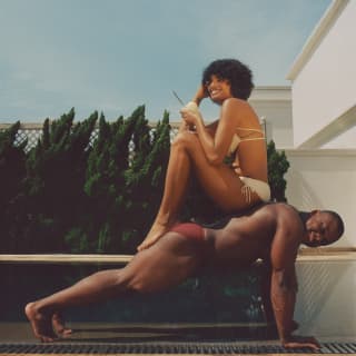 A woman with dark curly hair and a white bikini sits on the back of a muscly man doing push-ups in maroon gym trunks.