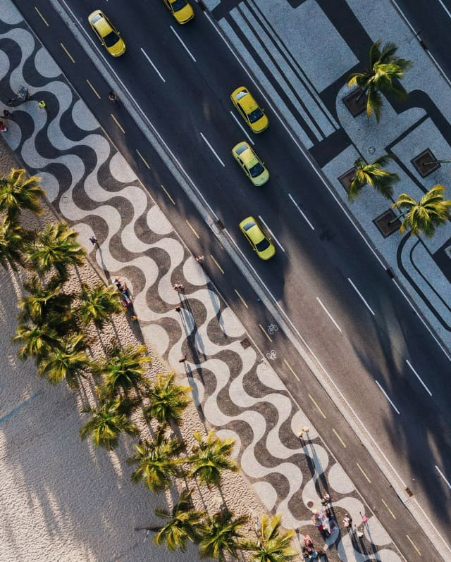 An aerial view shows the iconic black and white wavy lines of the Copacabana boardwalk, flanked by yellow cabs and palms