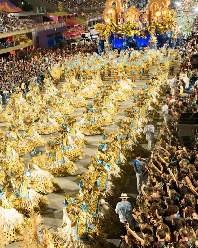 High-angle view of the Sambadrome Parade with dancers in straw-like costumes behind a blue and gold float, flanked by crowds.