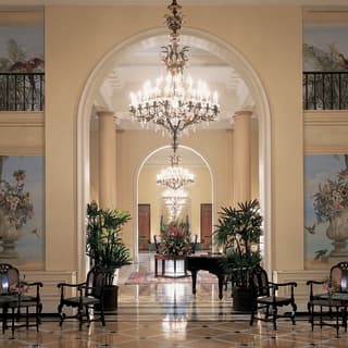A large chandelier hangs from a double height arch, illuminating marble floors and frescoed walls in a large elegant foyer