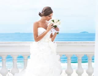 A bride in a cloud dress with sweetheart neckline looks at her posy and she stands on the balcony before the turquoise sea