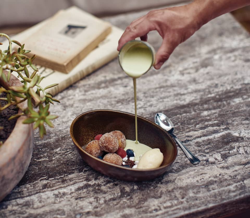 Close-up of custard being poured into an oval bowl of donut balls
