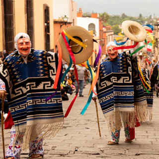 Three people parading in masks and blue traditionally-woven ponchos wave wide-brimmed hats teeming with rainbow ribbons.