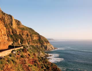 Ocean road curving around a sloping cliff beside a calm ocean at sunset