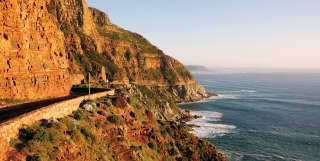Ocean road curving around a sloping cliff beside a calm ocean at sunset