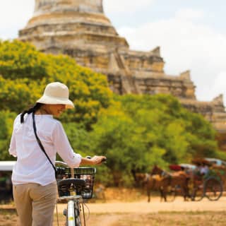 Lady holding a bicycle on a dusty road with a temple rising beyond