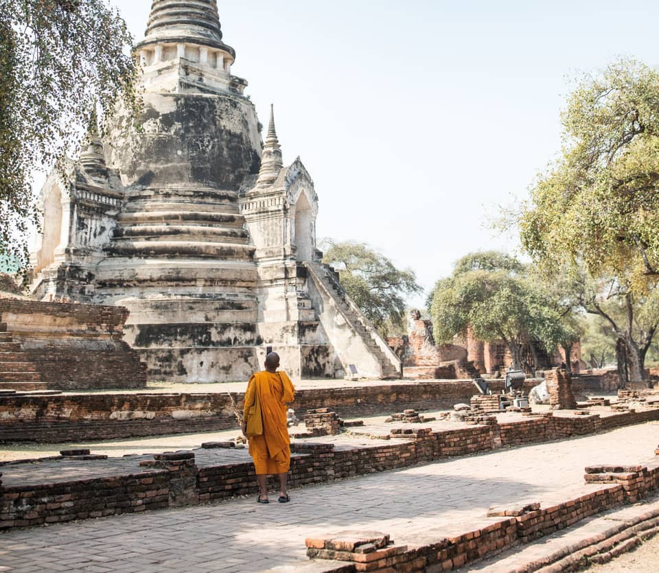 A monk in orange robes looks up at the conical peak of a stone monument in Ayutthaya's holy temple of Wat Phra Si Sanphet.