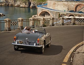 Dusk grey Italian classic soft-top car being driven on a coastal road