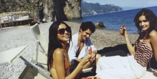 On sun loungers, a smiling man and two women with long dark hair, dressed to swim, share plates of cool sliced watermelon.