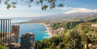 View from Capo Taormina south over the Ionian coast, with pretty seaside towns hemmed in by bright sea and steep green hills.