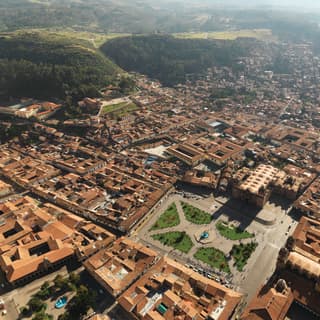 The open space and green gardens of the historic Plaza de Armas stands out in the red-roof cluster of Cusco, seen from above.